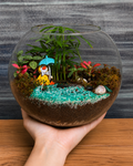 Donald Duck Terrarium from Planterie holded by a hand, a miniature glass terrarium with a Donald Duck figurine, pebbles, moss and a small pond arrangement to look like a duck setting, shown with care tips for indoor display in Indian homes and offices
