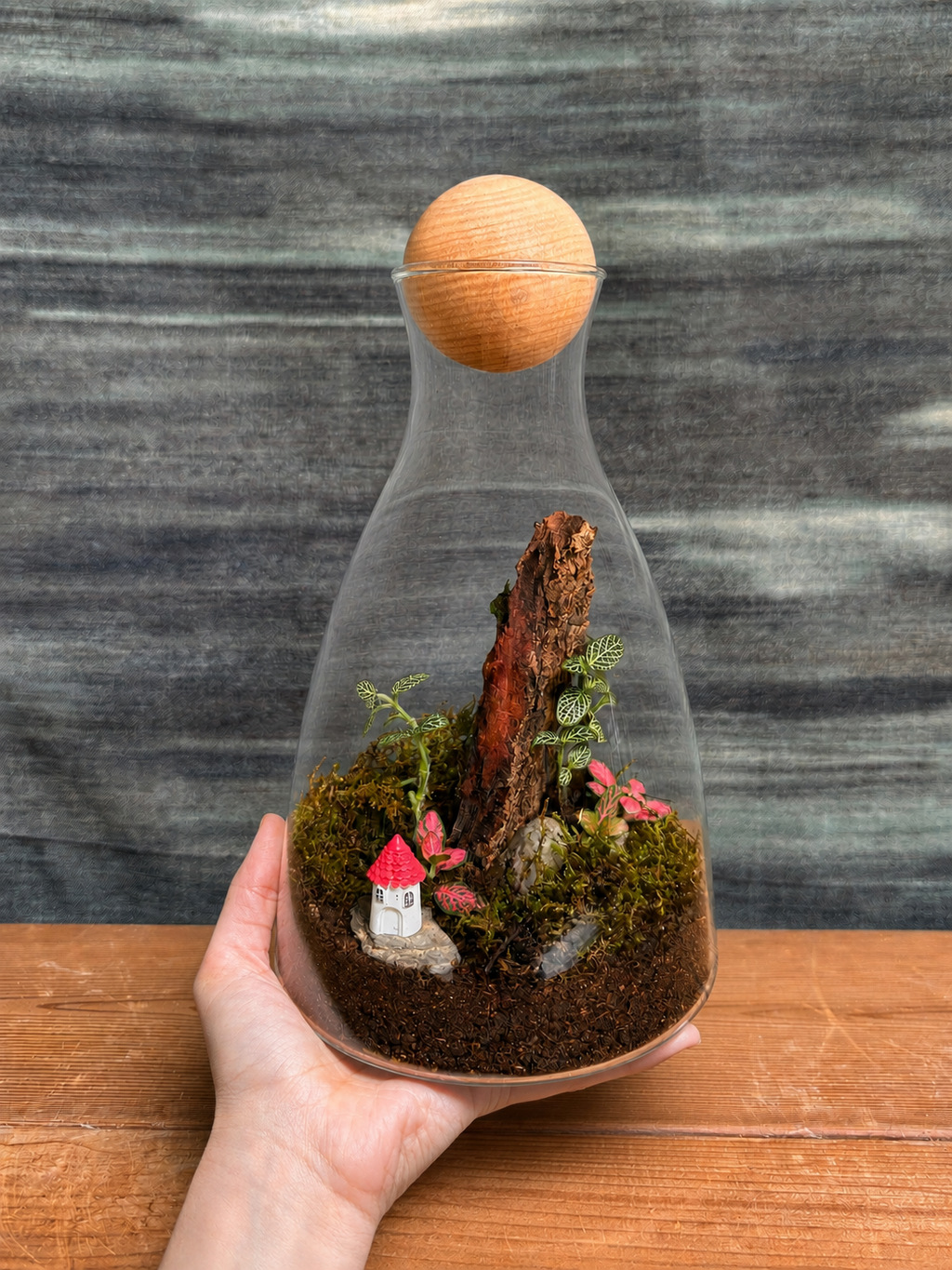 Forest Cottage Terrarium from Planterie holded by a hand, a miniature glass decanter terrarium with a tiny cottage figurine, moss, pebbles and small greenery arranged like a forest cottage scene, perfect as a compact desktop terrarium for work tables and shelves in Delhi homes and offices
