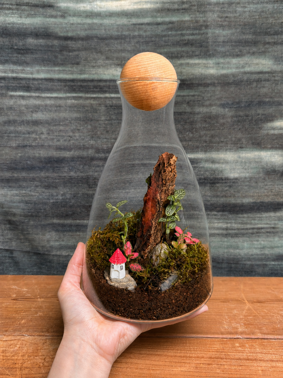 Forest Cottage Terrarium from Planterie holded by a hand, a miniature glass decanter terrarium with a tiny cottage figurine, moss, pebbles and small greenery arranged like a forest cottage scene, perfect as a compact desktop terrarium for work tables and shelves in Delhi homes and offices